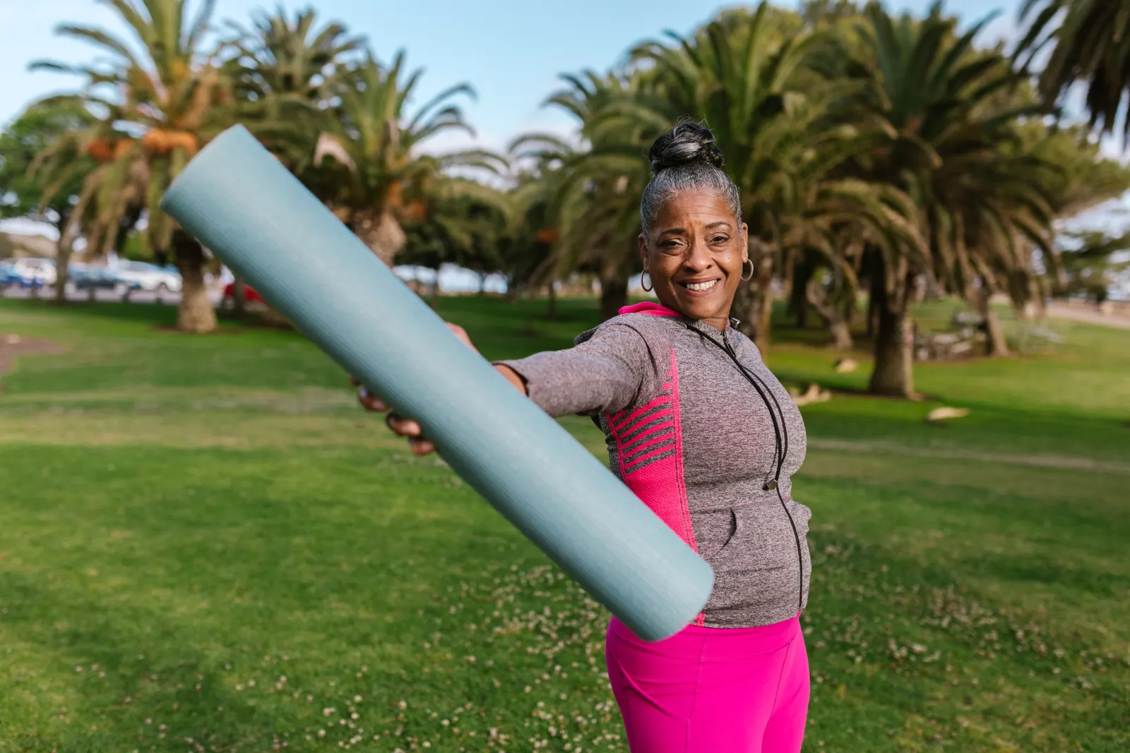 An active older woman smiling outdoors while holding an exercise mat.