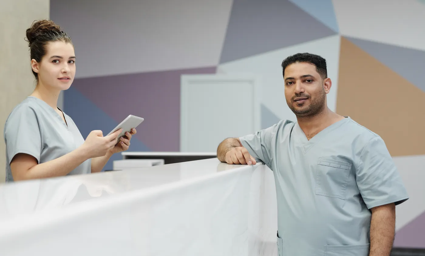 Two healthcare workers in scrubs at a clinic reception desk.