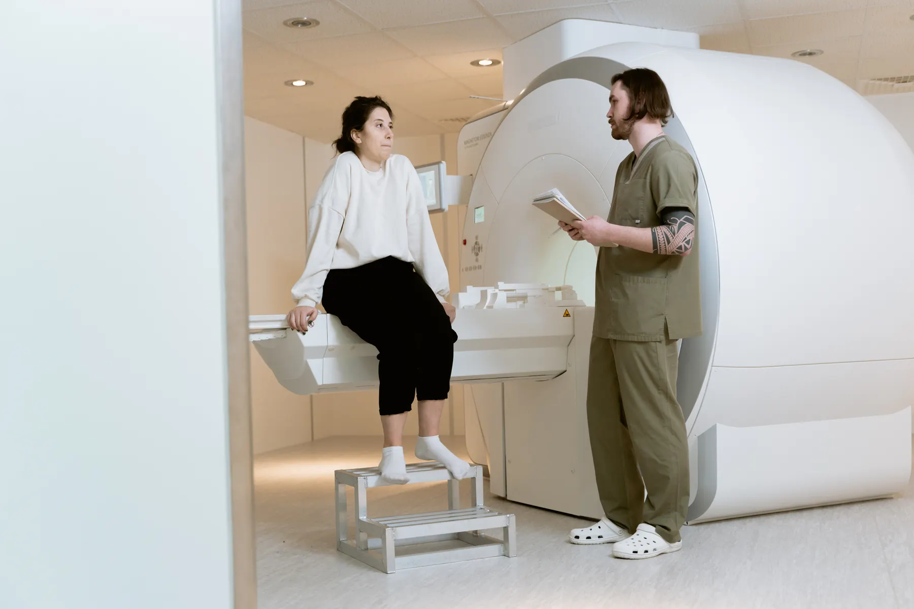 A patient sitting on an open scanner table with a radiographer beside her.
