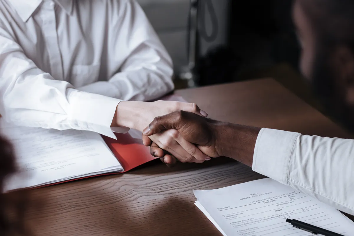 Two people shaking hands over signed documents at a desk.